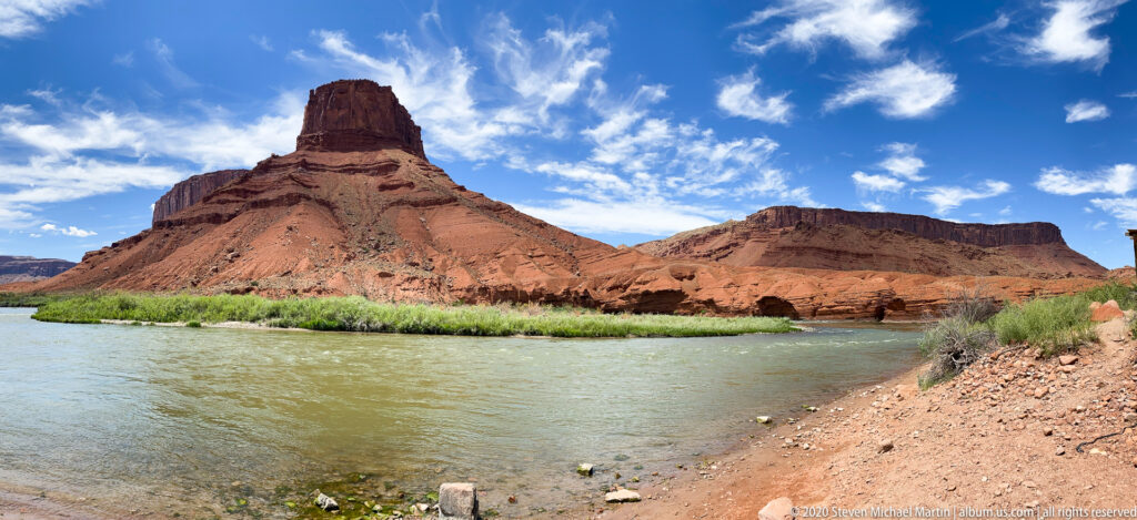 A sandstone spire and broad red formation rise beyond the Colorado River and a green riverbank.