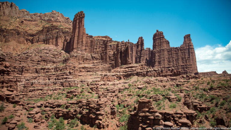 Tall sandstone pinnacles rise from layered red cliffs above a rocky slope with sparse shrubs.