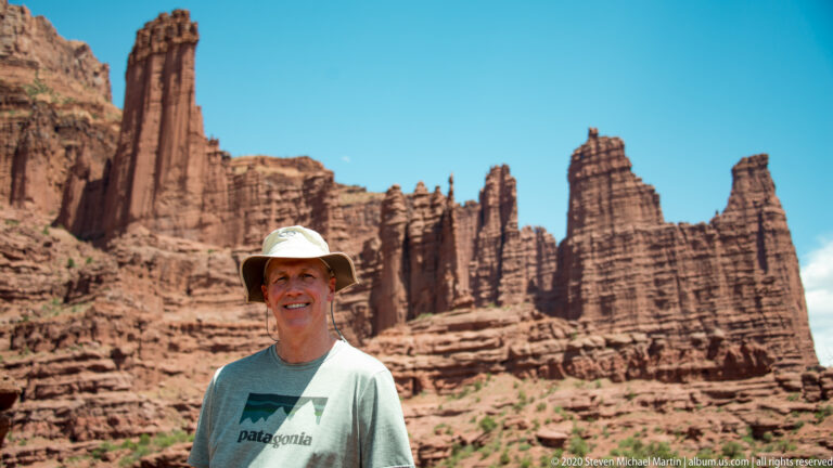 A visitor in a pale hat stands before towering sandstone pinnacles under a clear blue sky.