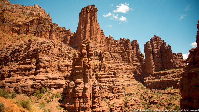 A tall fluted sandstone tower rises above clustered red spires against a bright sky.