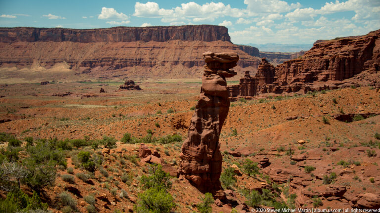 A rounded boulder balances on a narrow rock pillar above a broad red and green basin.