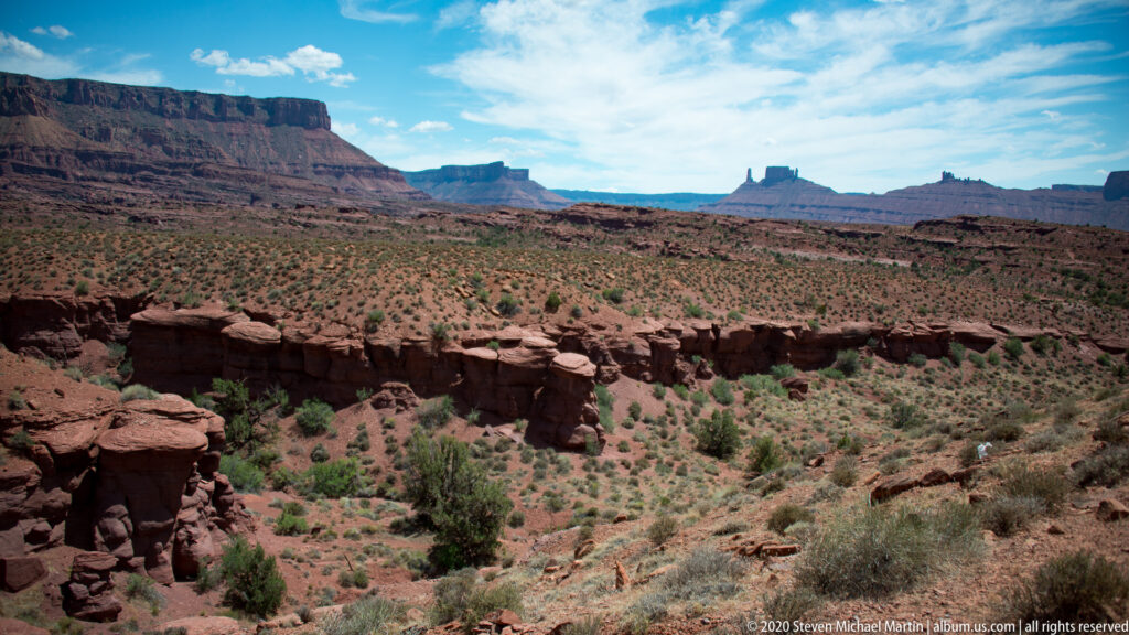 A cliff frames a broad shrub-covered basin leading toward distant sandstone towers.