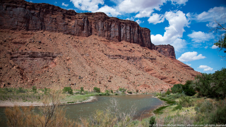 A green river runs below a high red sandstone cliff, with trees and gravel along the bank.