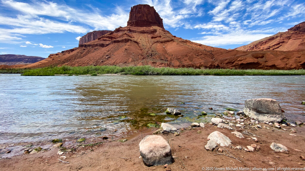 Shoreline stones and shallow water lead toward a distant sandstone spire and red formations across the river.