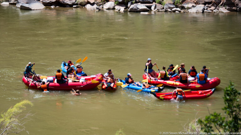Colorful rafts carrying passengers in life jackets float together beside a rocky riverbank.