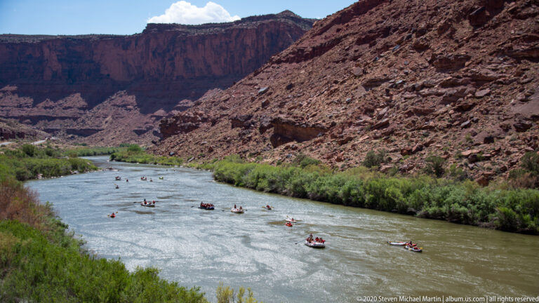 Several small rafts travel along a winding river bordered by green trees and steep red slopes.