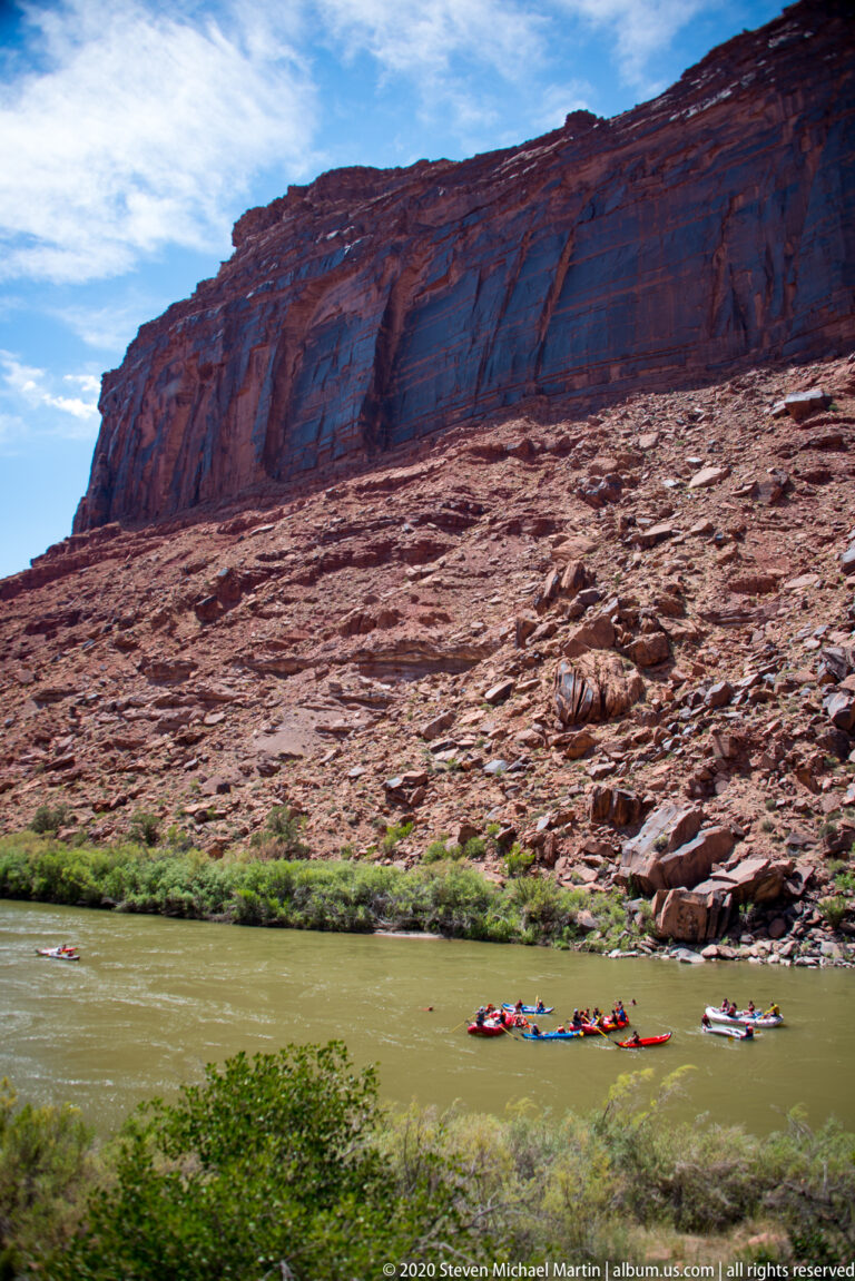 A line of small rafts crosses a river beneath a long red cliff with vertical seams.