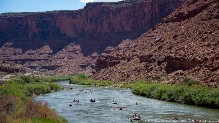 Several rafts follow a river bend between green banks and steep red canyon slopes.