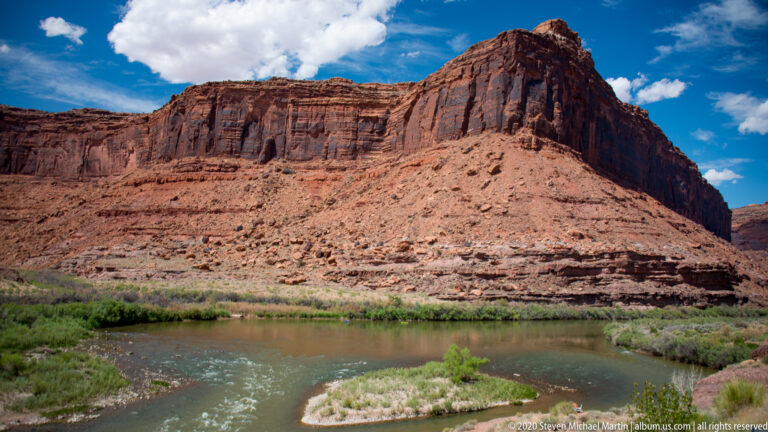 A vegetated river island lies below a broad layered sandstone bluff, with small rafts upstream.