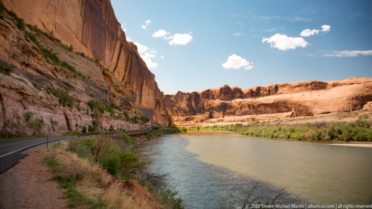 A paved road and green river curve together beneath tall sandstone walls and a blue sky.