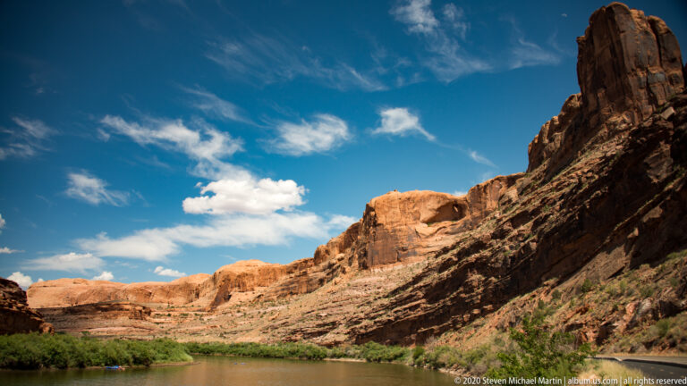 A shadowed cliff frames an orange sandstone wall across a river lined with green trees.