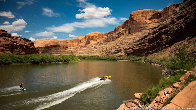 A raft leaves a white wake as a lone paddler glides beside it on a river below red cliffs.