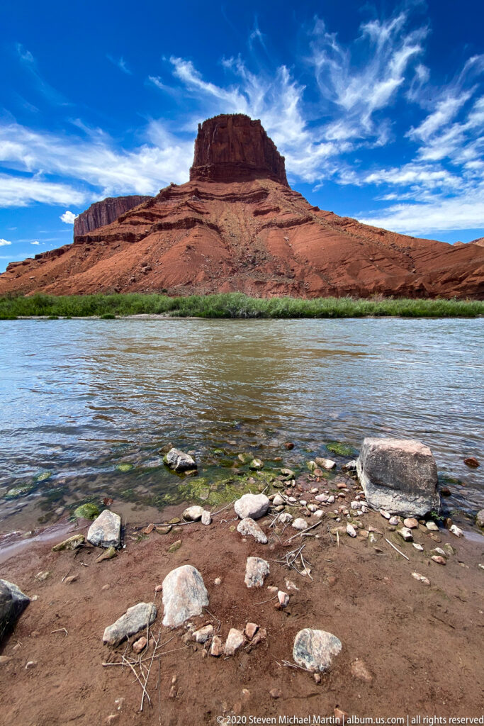 A broad red sandstone mesa rises across the Colorado River beyond a stony gravel shoreline.