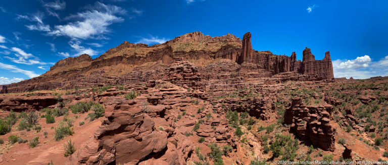 Several narrow sandstone fins project from a broad sloping red rock formation above sparse vegetation.