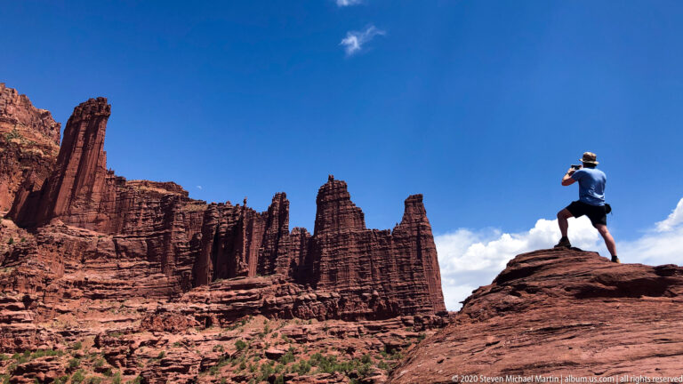 A visitor photographs tall sandstone fins while standing on a rocky perch above the surrounding terrain.