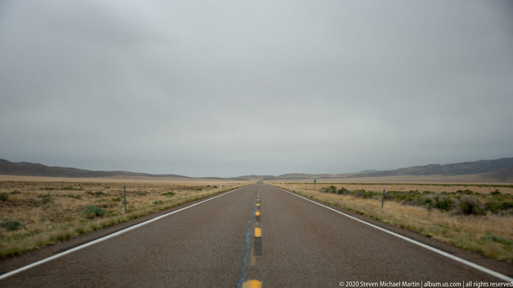 A straight two-lane highway crosses a dry basin toward low mountain ridges beneath a muted sky.