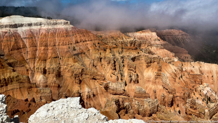 Layered orange cliffs and narrow hoodoos fill the amphitheater beneath a dark cloud and forested rim.