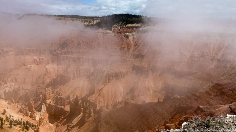 Mist veils the orange canyon walls below a forested rim, leaving only fragments of the amphitheater visible.