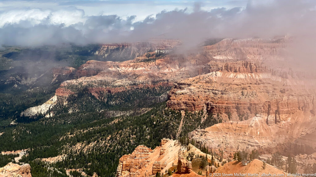 Cloud wisps cross a wide orange amphitheater bordered by dark forested ridges and pale rock bands.