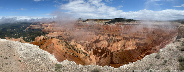 Rust-colored pinnacles and forested canyon slopes spread below an overlook under scattered clouds.