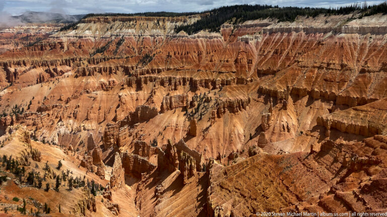 Rows of orange hoodoos and shadowed ravines fill the basin below a pale, forested rim.