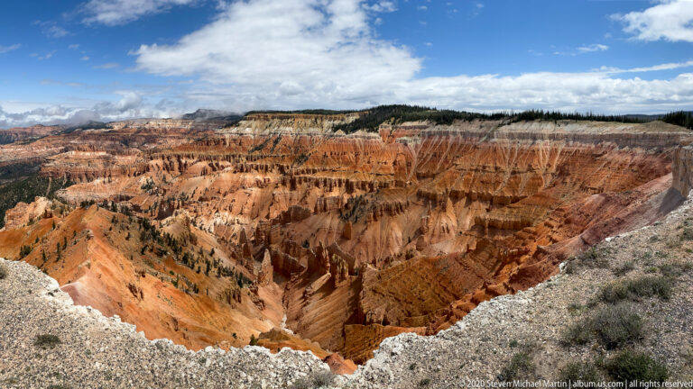 Pale broken rock frames an open orange amphitheater beneath a forested plateau and scattered clouds.