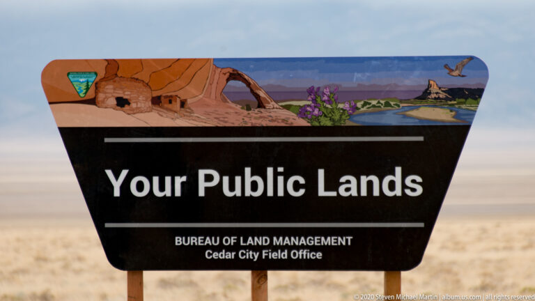 A Bureau of Land Management sign reading “Your Public Lands” stands before dry grassland and distant hills.