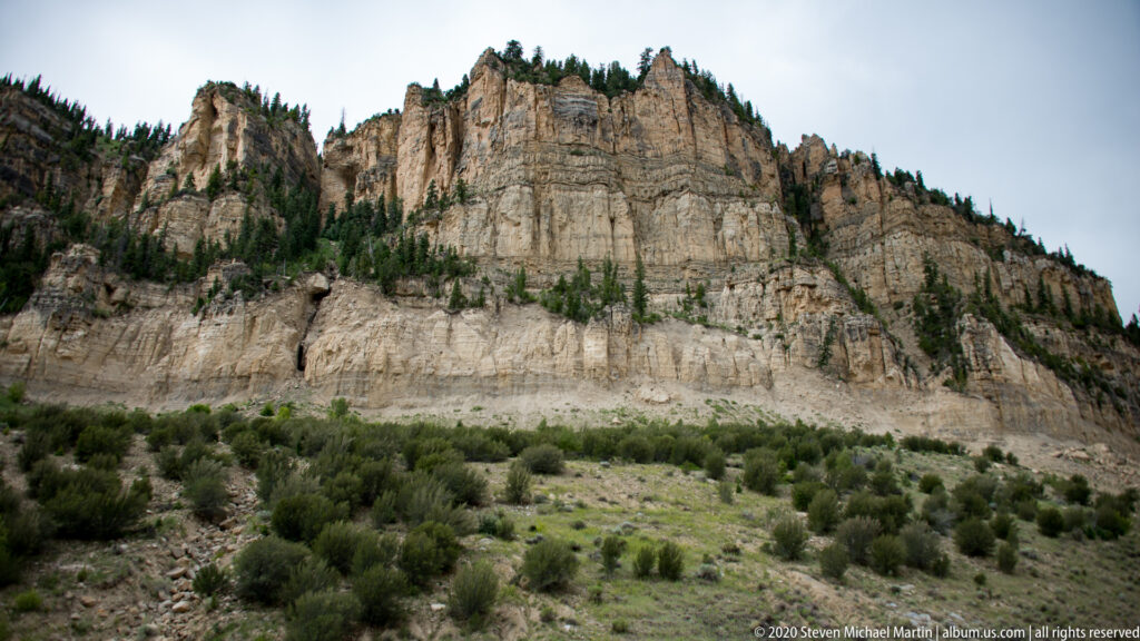 A pale stratified cliff rises above shrubs and conifers beneath a gray sky.