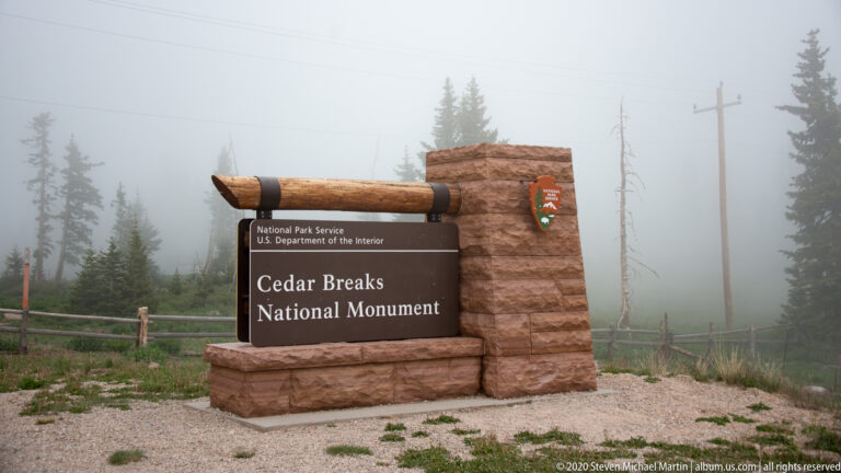 A Cedar Breaks National Monument entrance sign stands on a stone base beside a fence in fog.