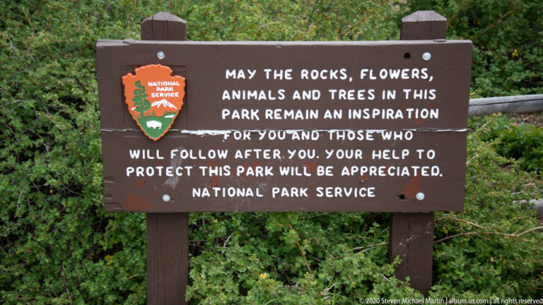 A National Park Service sign among green foliage asks visitors to help protect the park’s natural features.