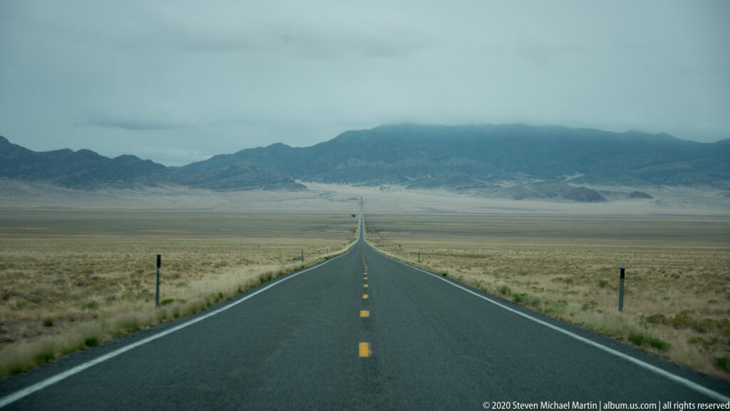 A highway descends across a broad grassland basin toward hazy mountain ridges beneath low clouds.