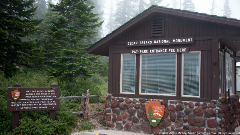 A Cedar Breaks entrance booth and National Park Service sign stand beside a wooden fence among conifers.