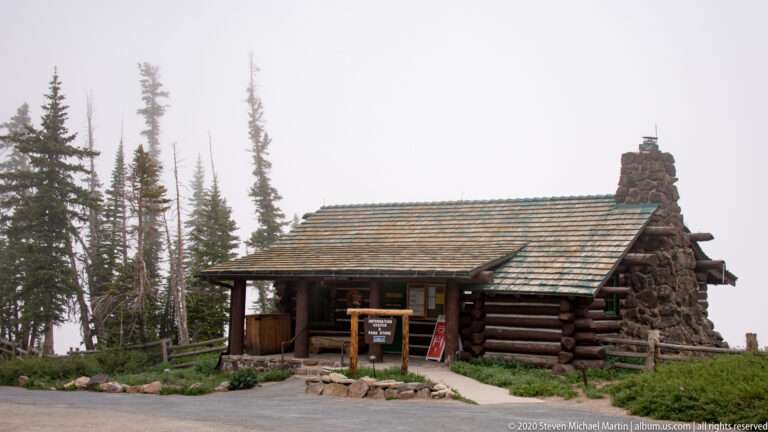 A log visitor center with a broad shingled roof and stone chimney stands among conifers.