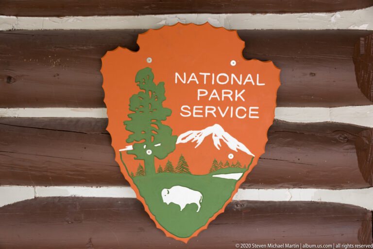 A National Park Service arrowhead plaque is mounted on a horizontal log wall.