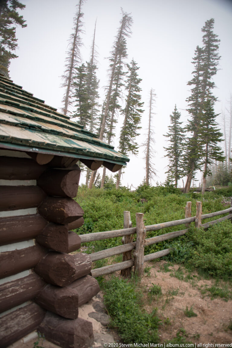 A log building corner and roof border a split-rail fence beside a misty conifer clearing.