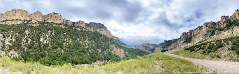 A curving road follows a forested canyon beneath steep pale cliffs and broken clouds.