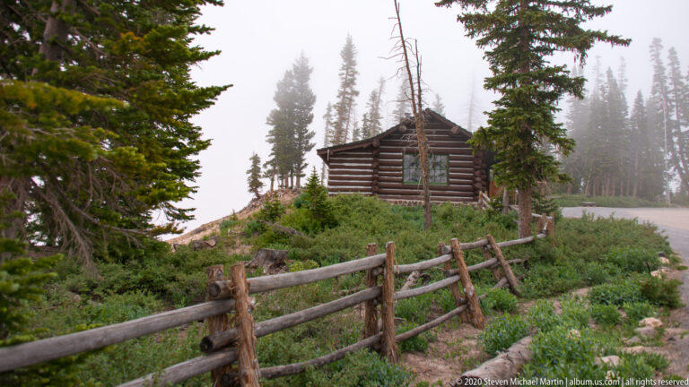 A log building stands behind a zigzag timber fence among conifers beside a foggy road.