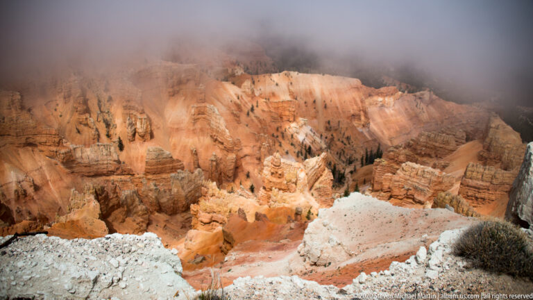 Orange and cream hoodoos descend into a broad amphitheater beneath low fog.