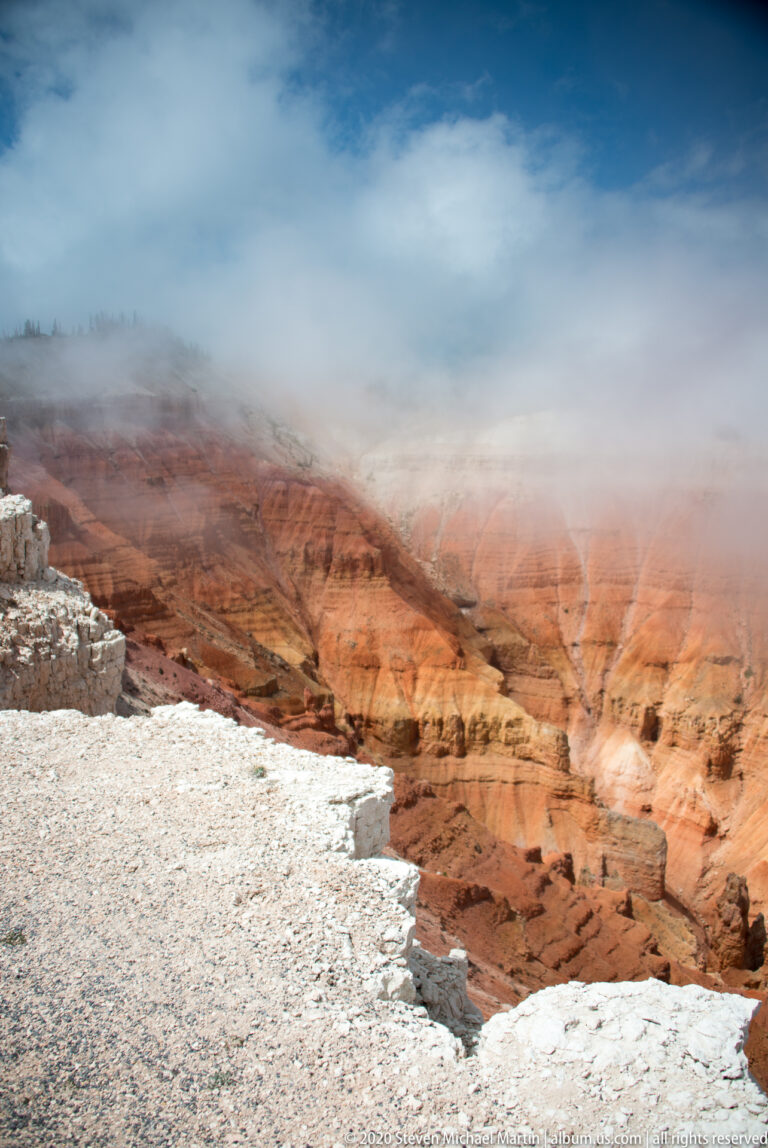 A pale rim ledge overlooks red hoodoos and layered cliffs partly hidden by low cloud.