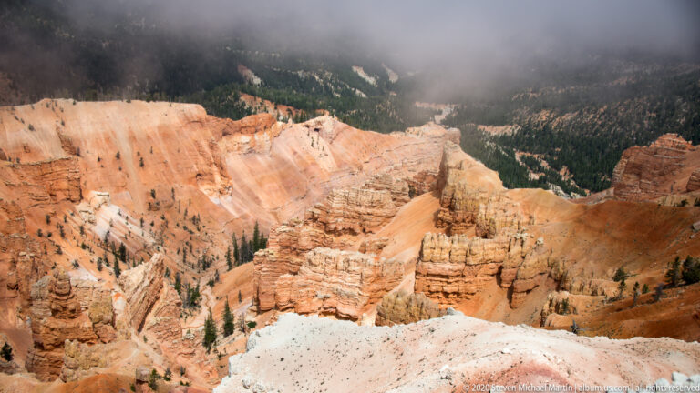Orange hoodoos descend toward a dark forest beneath drifting mist along the upper rim.