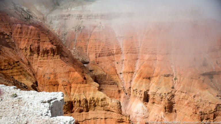 A pale rim shelf overlooks orange hoodoos and cliffs partly obscured by drifting cloud.