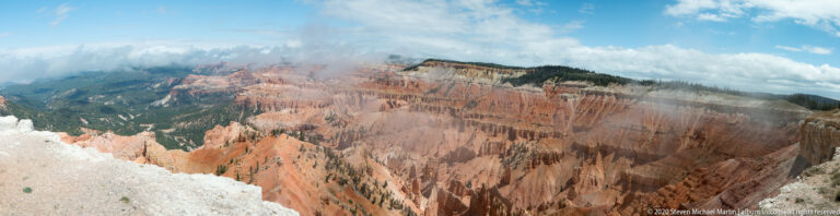 Clouds cross the rim above orange hoodoos and dark forest, with a pale ledge in the foreground.