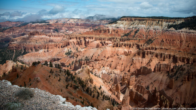A rough pale rim overlooks rows of orange hoodoos and forest fading into a hazy plateau.