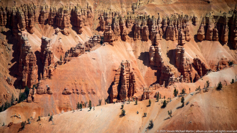 Rows of narrow orange and cream hoodoos stand above a pale slope with scattered trees.