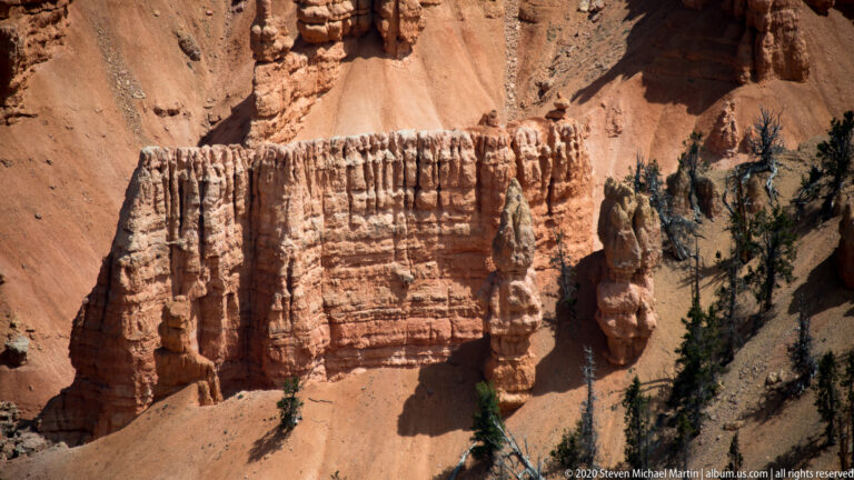 Horizontal rock bands are broken by vertical fins, with dark trees on the slope at right.