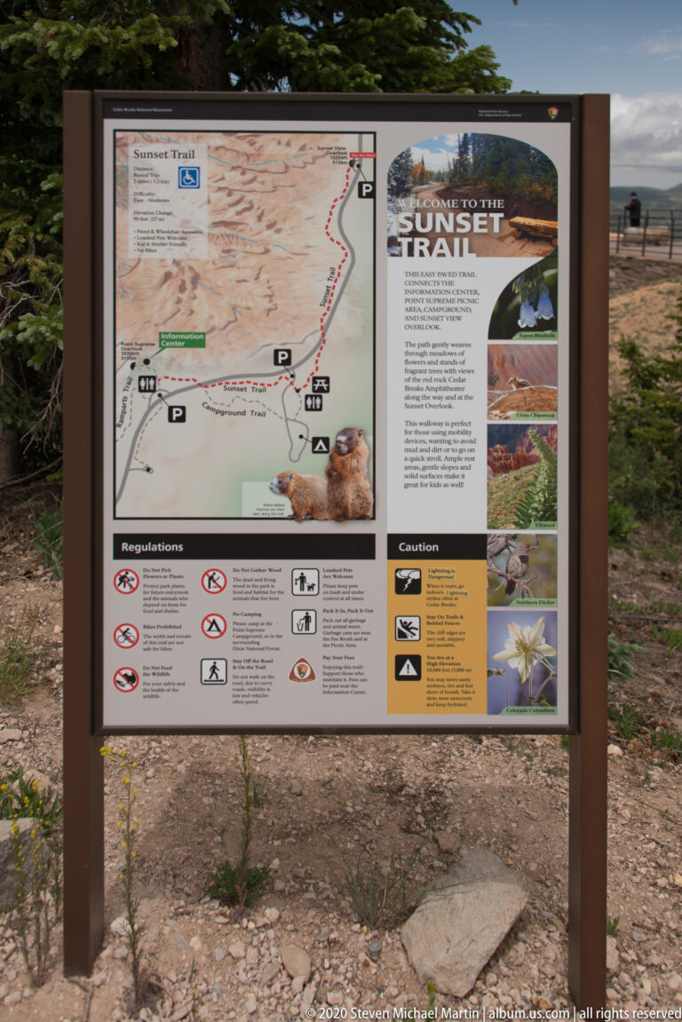 A framed visitor sign displays a trail map, regulations, and a Sunset Trail panel in a shrub-lined clearing.