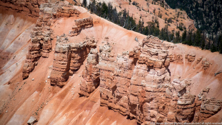 Orange banded columns rise from a pale ridge, with conifers on the darker slope behind.