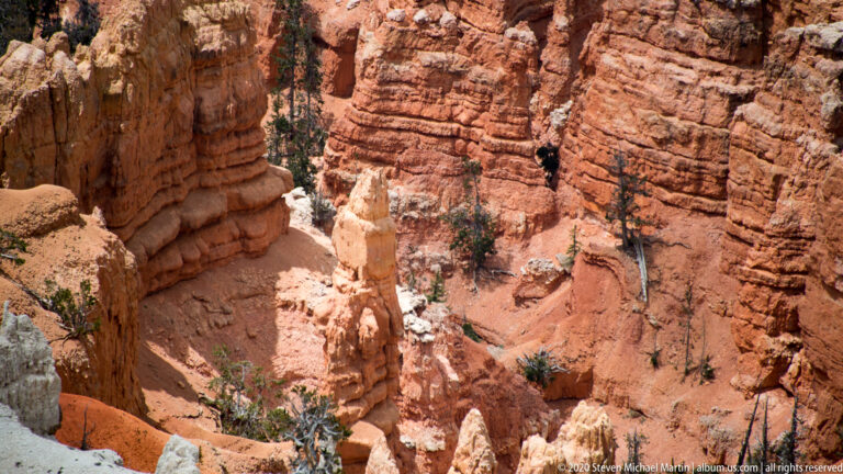 A dark cleft separates steep orange and cream columns, with a small tree on a ledge.