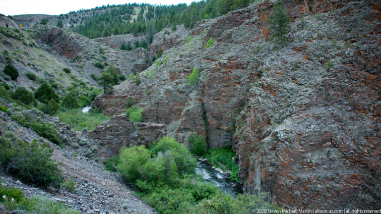 A small stream curves through a green-lined ravine between steep gray and reddish rock slopes.