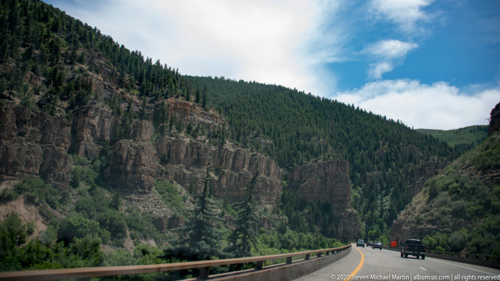 A paved highway and guardrail curve through a forested canyon beneath steep cliffs and a patch of blue sky.